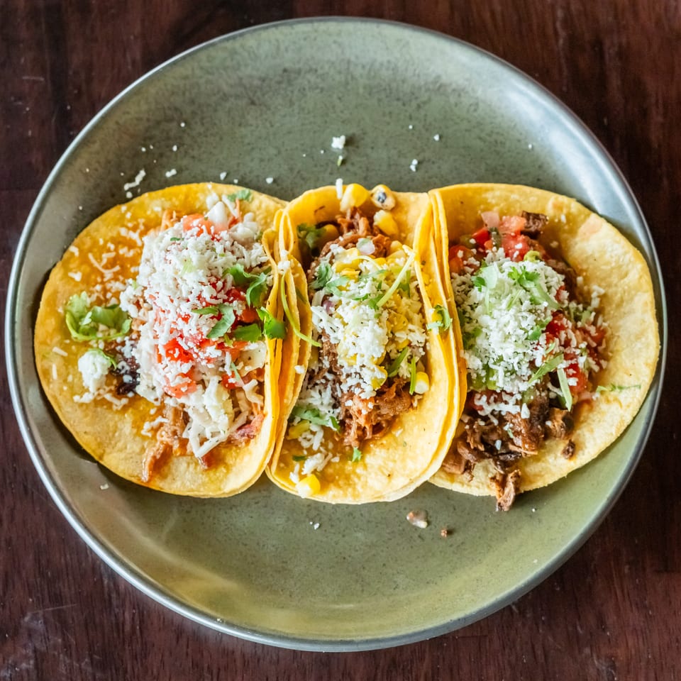 Three vibrant tacos with various fillings, cheese, and cilantro on a green plate.