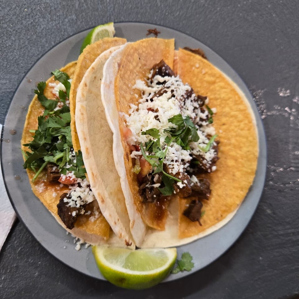 Plate of three street tacos with meat, cotija cheese, cilantro, and lime on a dark table.