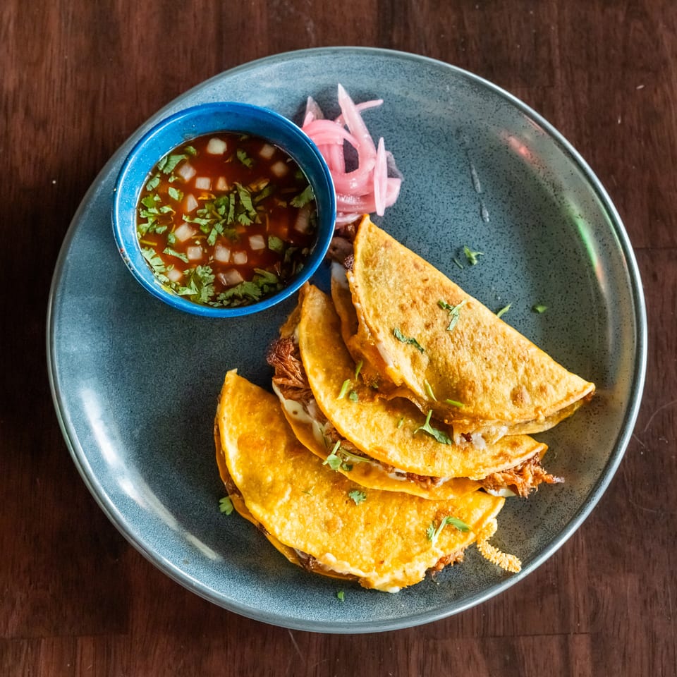 Three golden-brown birria tacos with melted cheese and meat, served with consommé and pickled onions.