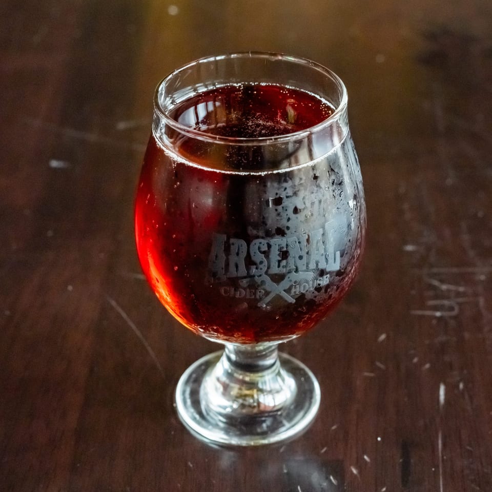 A glass of dark red Arsenal Cider House cider with condensation sits on a dark wooden table.