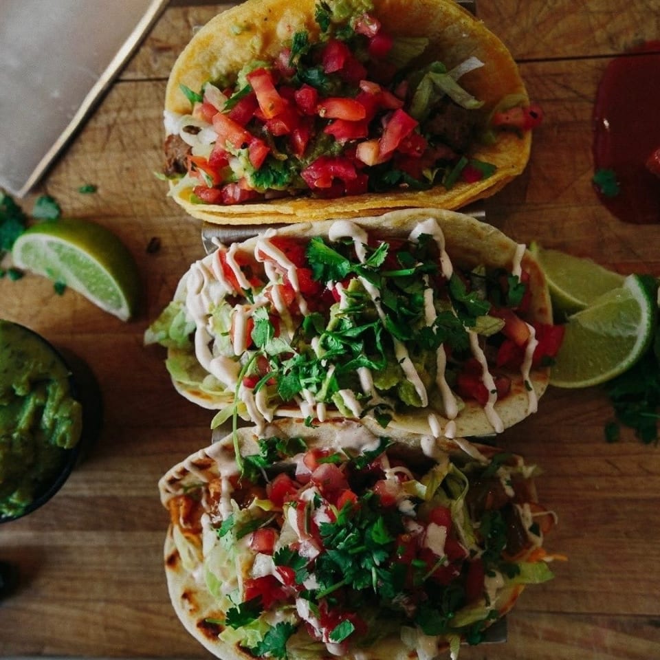 Three loaded tacos topped with chopped tomatoes, cilantro, lettuce, and sauce on a wooden board with guacamole, lime wedges, and hot sauce.