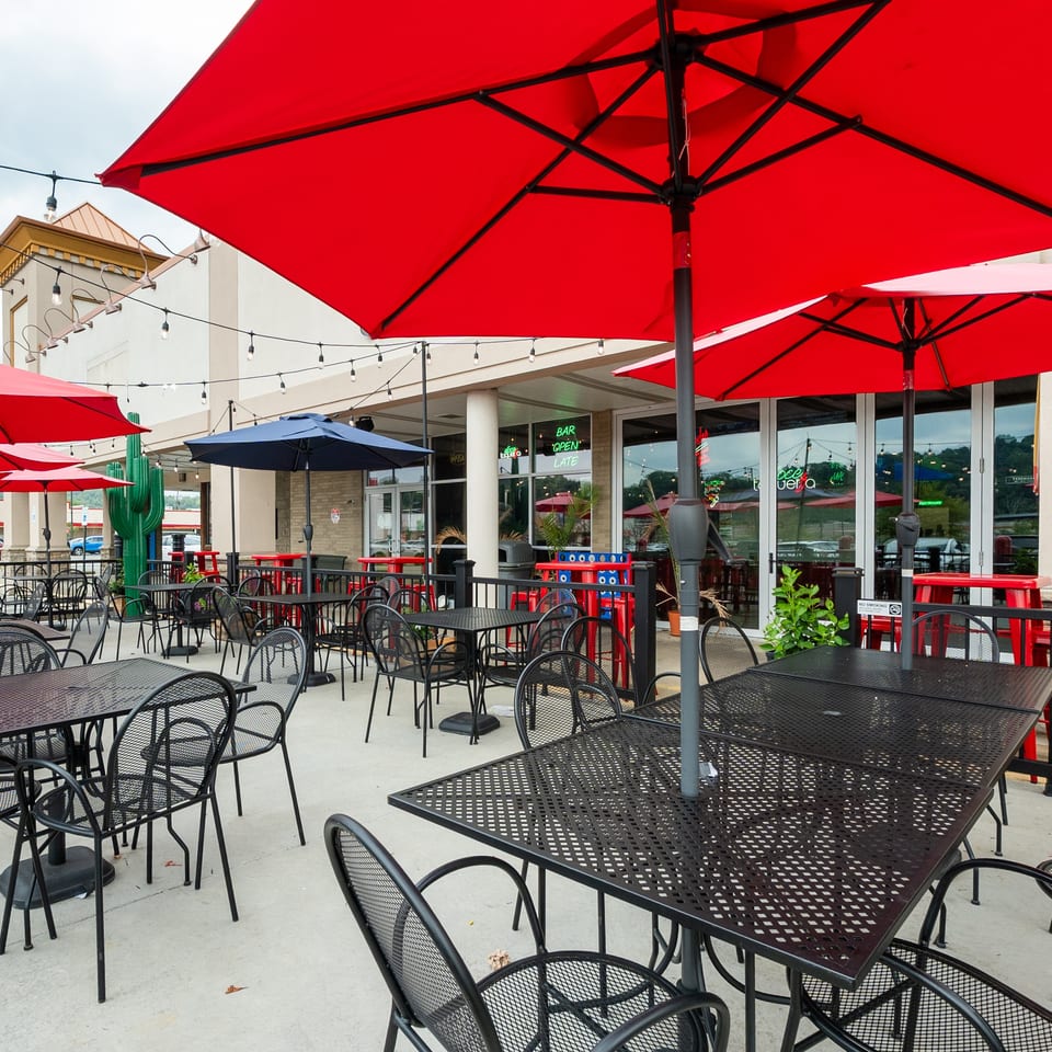 Outdoor patio with black metal tables and chairs, red and blue umbrellas, and string lights.