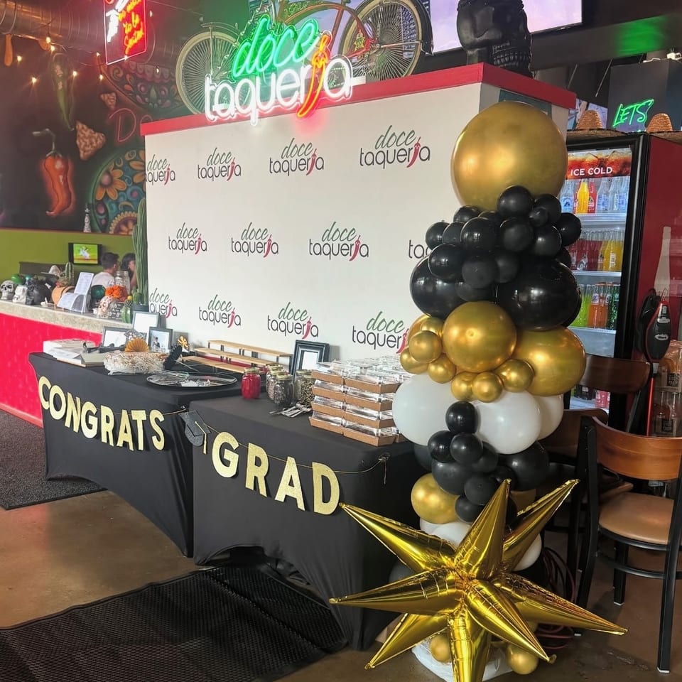 Graduation celebration display at Doce Taqueria featuring branded backdrop, dessert table, balloon column, and gold star decorations.