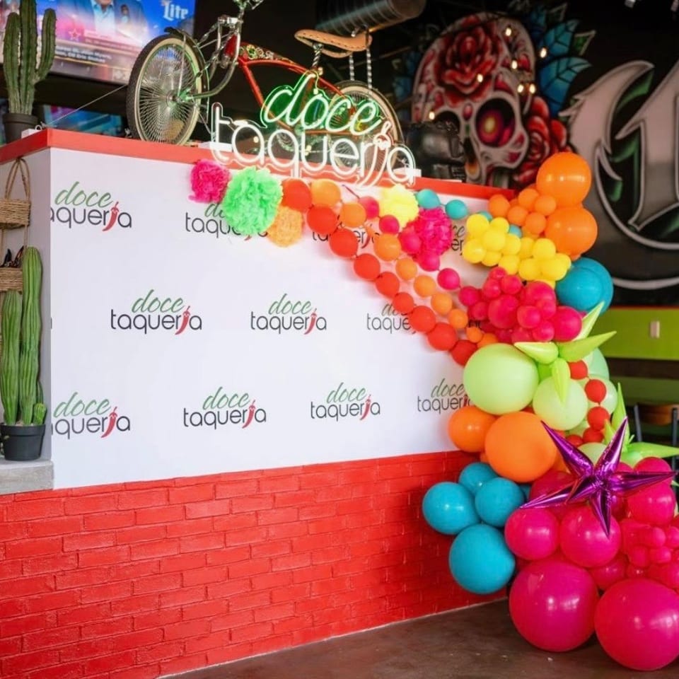 Colorful balloon arrangement decorates the counter at doce taqueria, surrounded by cactus plants, bicycles, neon signage, and vibrant mural art.