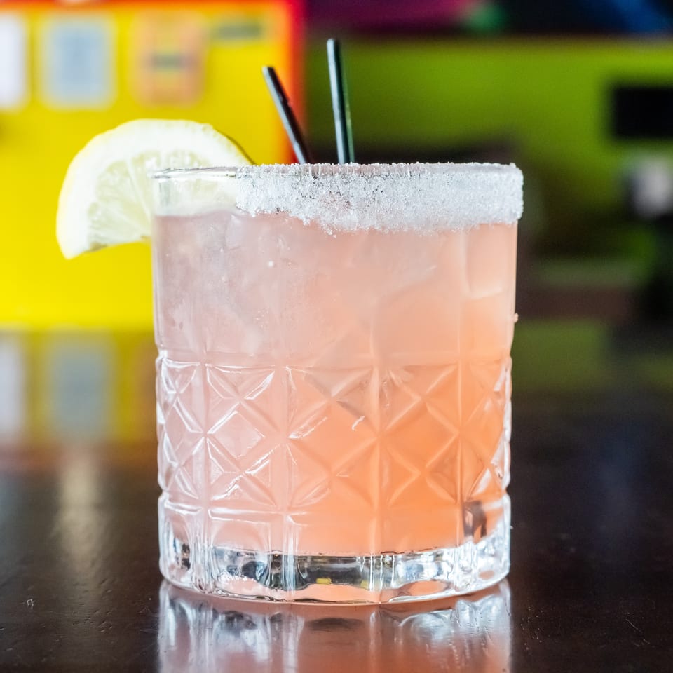 A pink cocktail in a textured glass with a salted rim, lemon slice, and two straws.