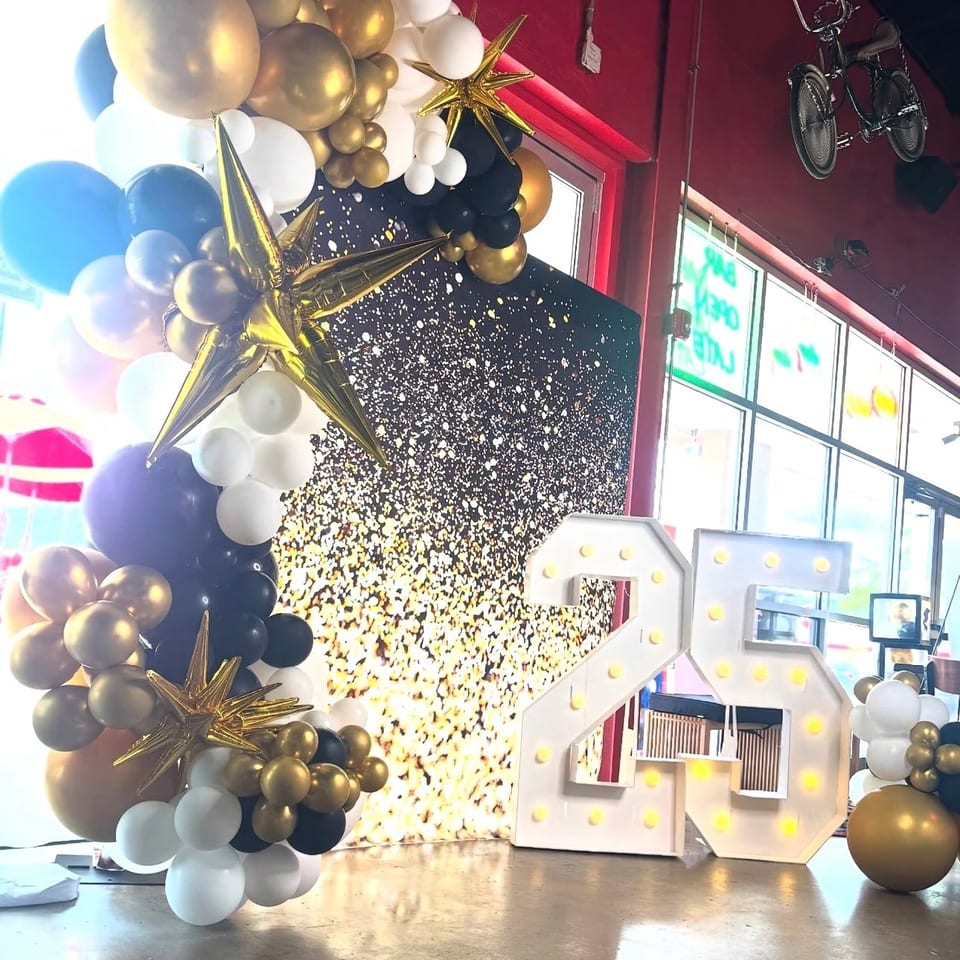 Festive restaurant display with gold, white, and navy balloon clusters, metallic stars, marquee numbers, and a glittering backdrop.
