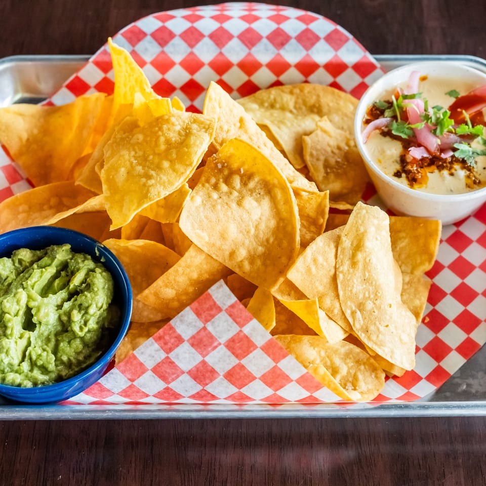 A tray of tortilla chips with guacamole and queso dip, garnished with pickled onions and cilantro.