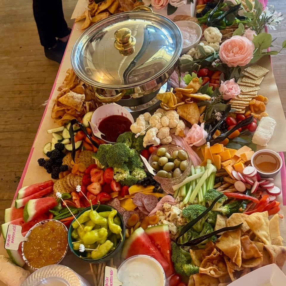 A lavish grazing table features covered chafing dishes, assorted cheeses, meats, fruits, vegetables, dips, crackers, chips, and floral arrangements.