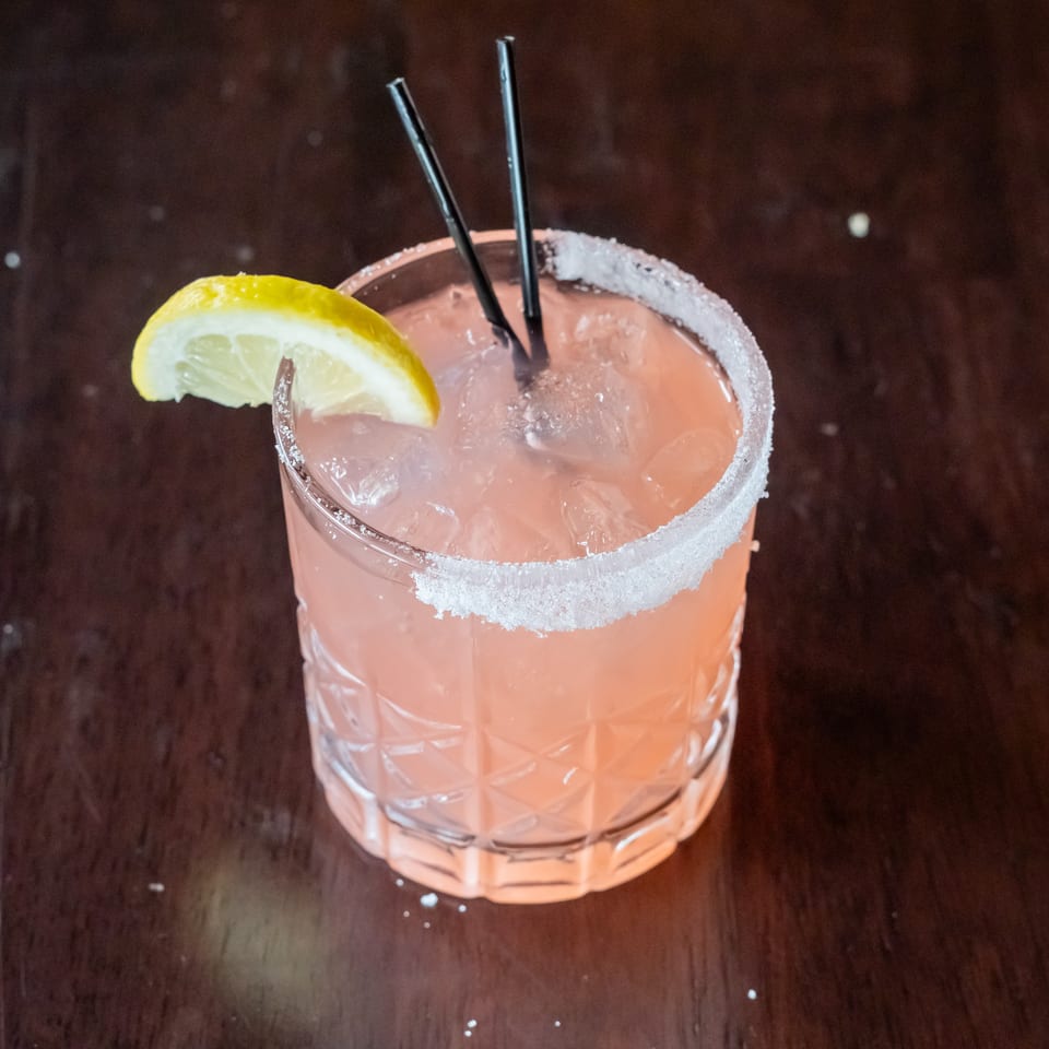 Pink cocktail with ice, a lemon wedge, and a sugar-rimmed glass on a dark wooden table.