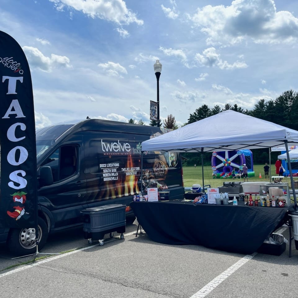 Taco food truck and tented serving booth at an outdoor festival, with inflatables and trees in the background.