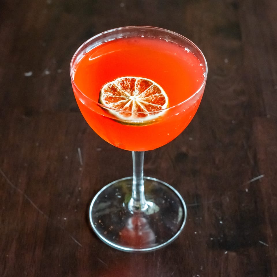 A vibrant red cocktail in a coupe glass, garnished with a dried lime slice, on a dark wooden table.