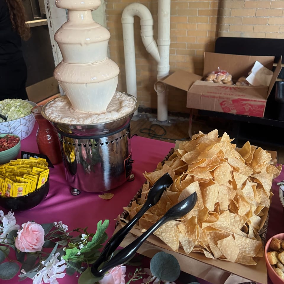 Buffet table with a decorative beverage dispenser, tortilla chips, condiments, and pink floral decorations against a brick wall.